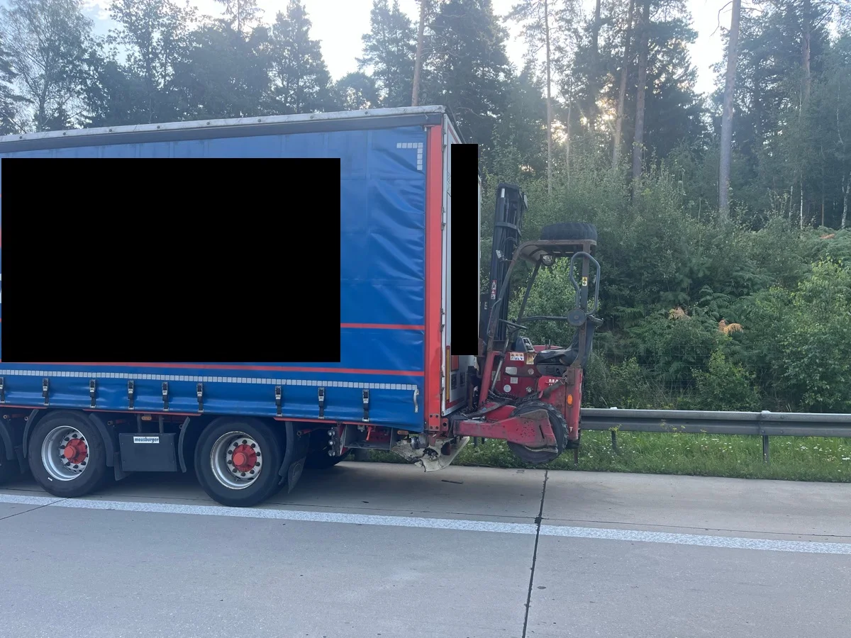 Ein blauer Lkw mit rotem Anhänger steht auf einer Autobahn. Im Hintergrund ist ein Wald zu sehen.
