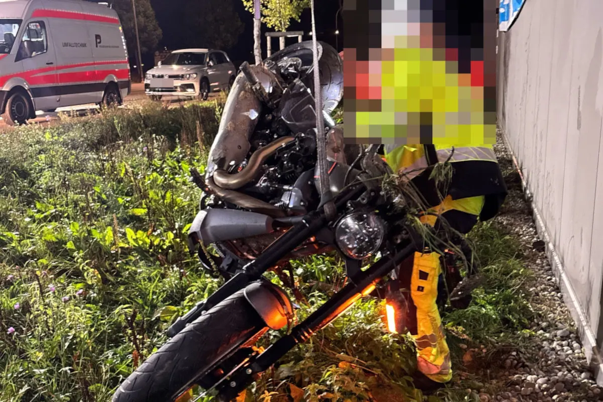 Ein stark beschädigtes Motorrad liegt bei Nacht auf einer Wiese neben der Straße. Eine Person in Warnkleidung ist daneben zu sehen.