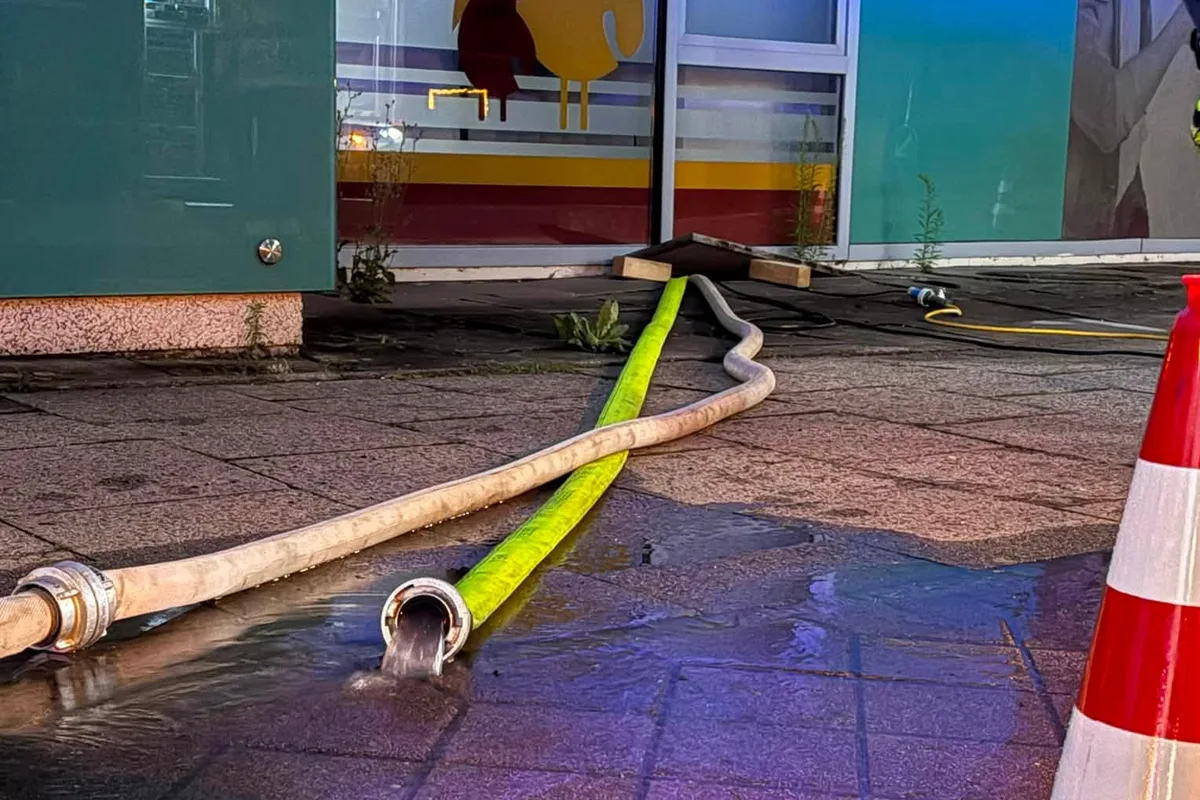 Zwei Feuerwehrschläuche liegen auf einem nassen Gehweg, aus einem fließt Wasser. Im Hintergrund ist ein Gebäude mit verschlossener Tür zu sehen.