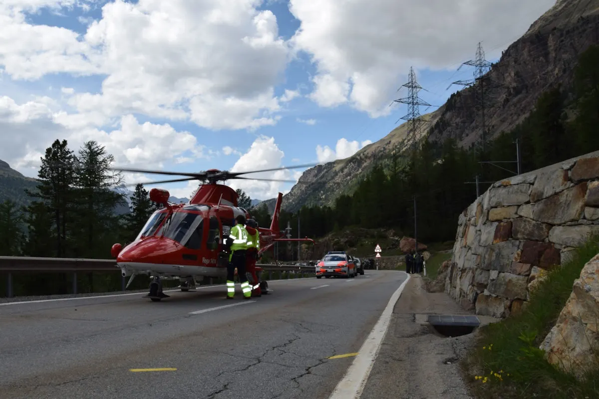 Ein roter Rettungshubschrauber steht auf einer Bergstraße. Helfer in gelben Westen sind in der Nähe. Ein Polizeiauto parkt dahinter.