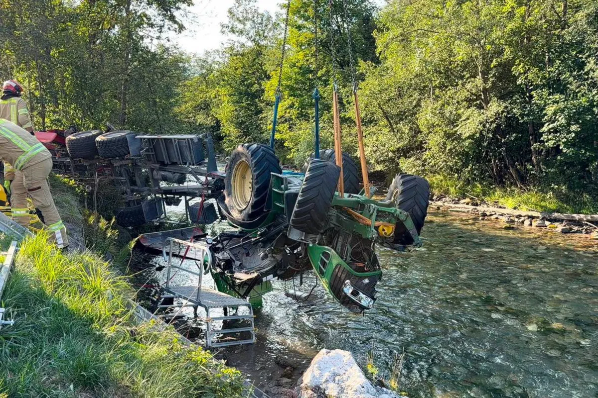 Der umgestürzte Traktor wird mit Seilen aus einem flachen Fluss gehoben, während ein Feuerwehrmann auf der Böschung steht.