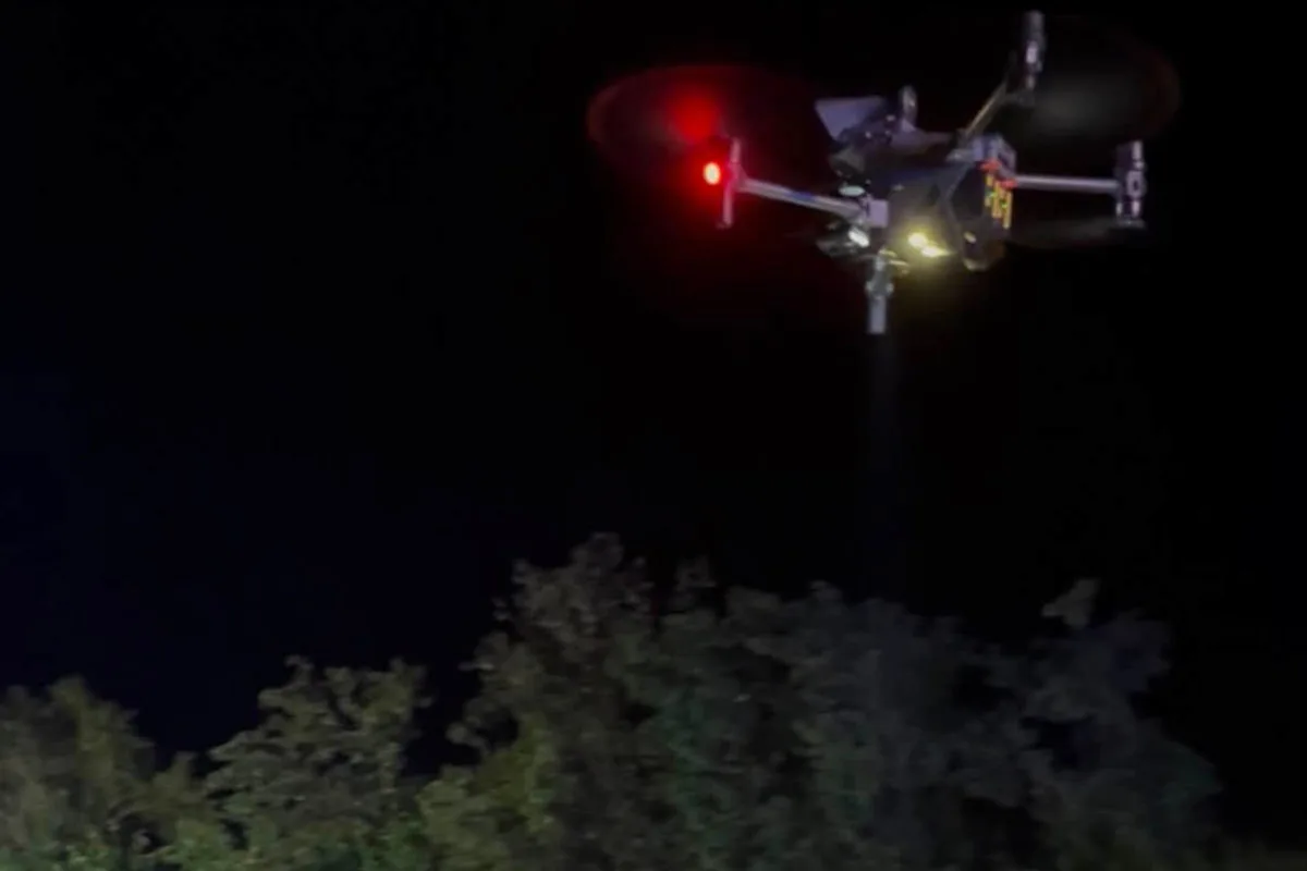 Eine Drohne mit roten und weißen Lichtern schwebt bei Nacht über Baumkronen. Der Himmel ist dunkel und die Bäume sind im unteren Bereich des Bildes sichtbar.