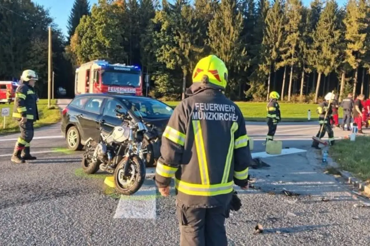 Mehrere Feuerwehrmänner in Schutzkleidung stehen an einer Unfallstelle mit einem Motorrad, einem Auto und einem Feuerwehrfahrzeug im Hintergrund.