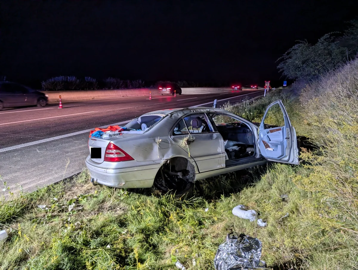 Ein silbernes Auto liegt schwer beschädigt am Straßenrand. Die Türen sind offen, und es ist Nacht.