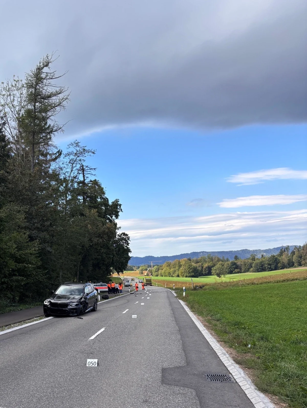Ein schwarzes Auto steht beschädigt am Straßenrand einer Landstraße, während mehrere Personen in orangenen Westen nahe der Leitplanke stehen. Am Horizont sind Felder und bewaldete Hügel zu sehen.
