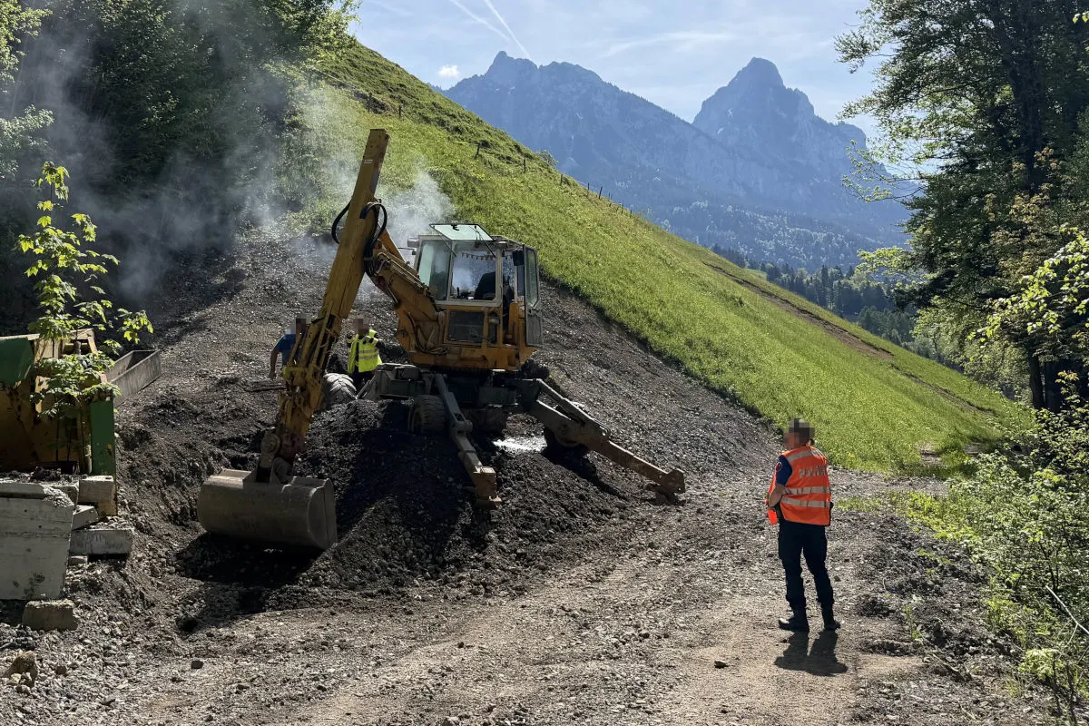 Ein Bauarbeiter in einer orangenen Warnweste steht neben einem gelben Bagger auf einer geneigten Bergbaustelle. Im Hintergrund sind Berge und Bäume sichtbar.