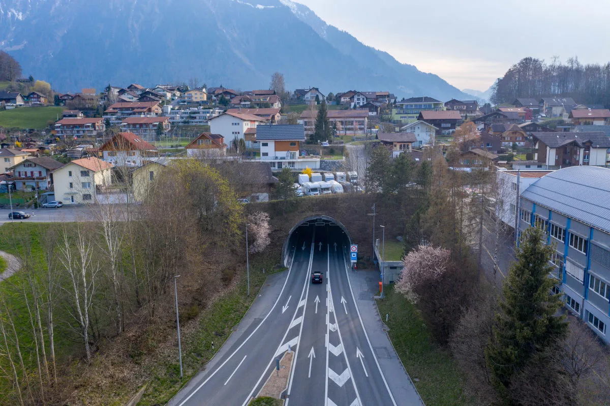 Ein Tunnel liegt am Ausgang eines Dorfs in bergiger Landschaft. Ein Auto fährt in den Tunnel und es ist früher Abend.