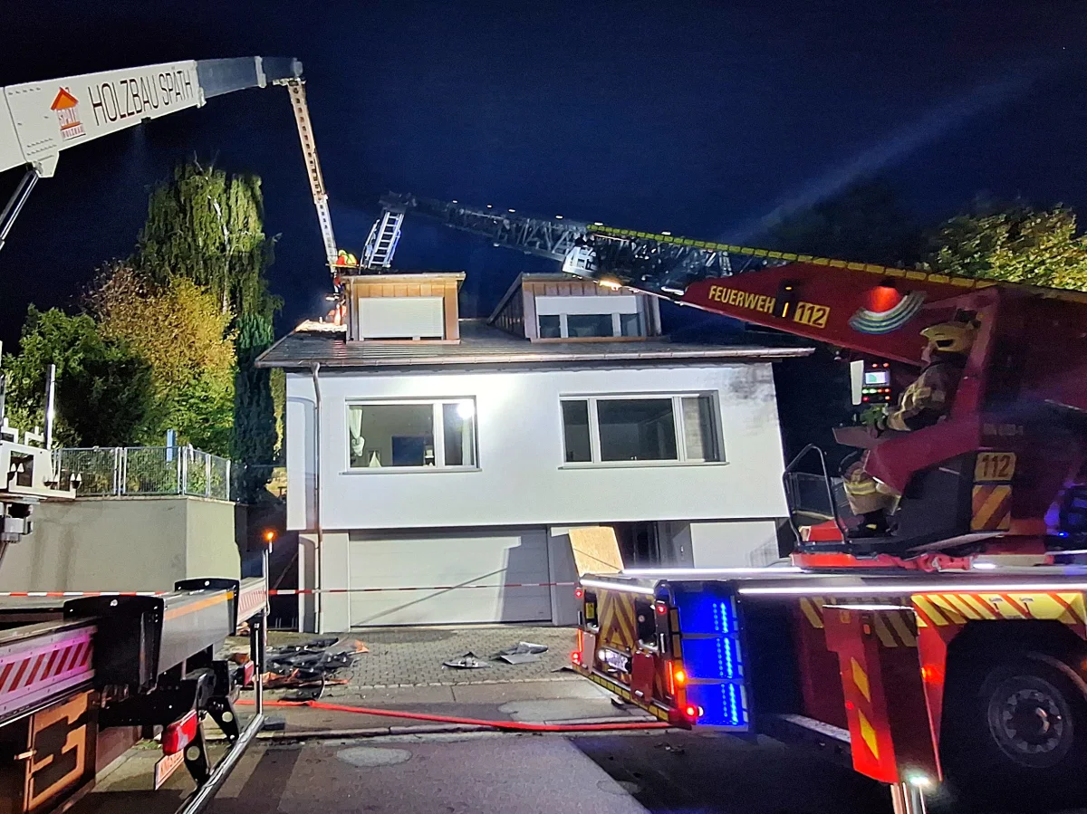 Ein Feuerwehrfahrzeug mit ausgefahrener Drehleiter bekämpft bei Nacht einen Brand auf dem Dach eines Hauses. Einige Feuerwehrleute sind auf der Drehleiter zu sehen, während der Bereich abgesperrt ist.
