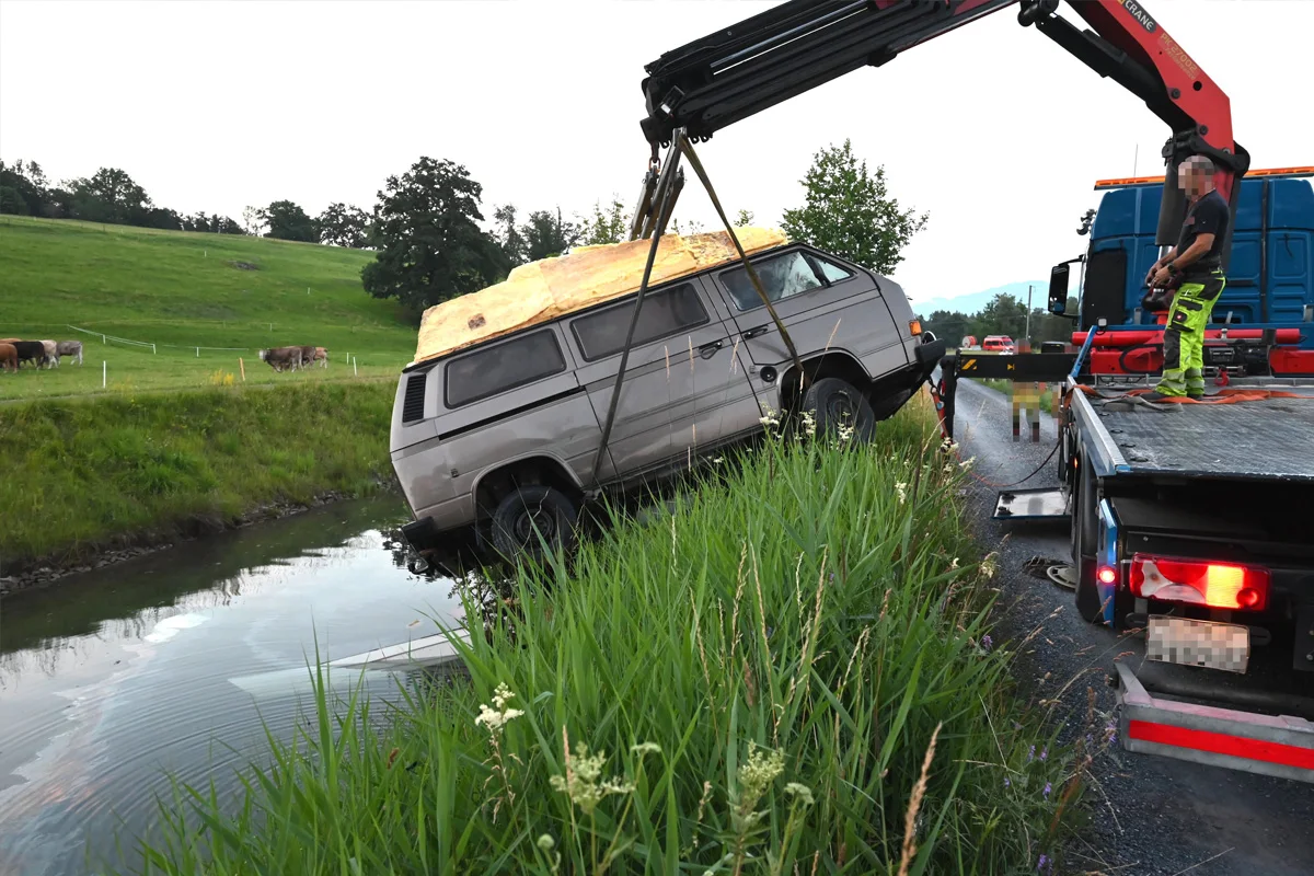Ein Kleinbus wird von einem Kran aus einem Wassergraben gehoben. Im Hintergrund sind eine Weide und Kühe zu sehen.
