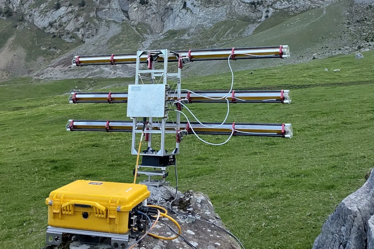 Ein Wetterradar-Gerät mit mehreren Antennen ist auf einem Felsen installiert, daneben steht ein gelber Koffer. Das Feld ist grün und im Hintergrund sind Felsen zu sehen.