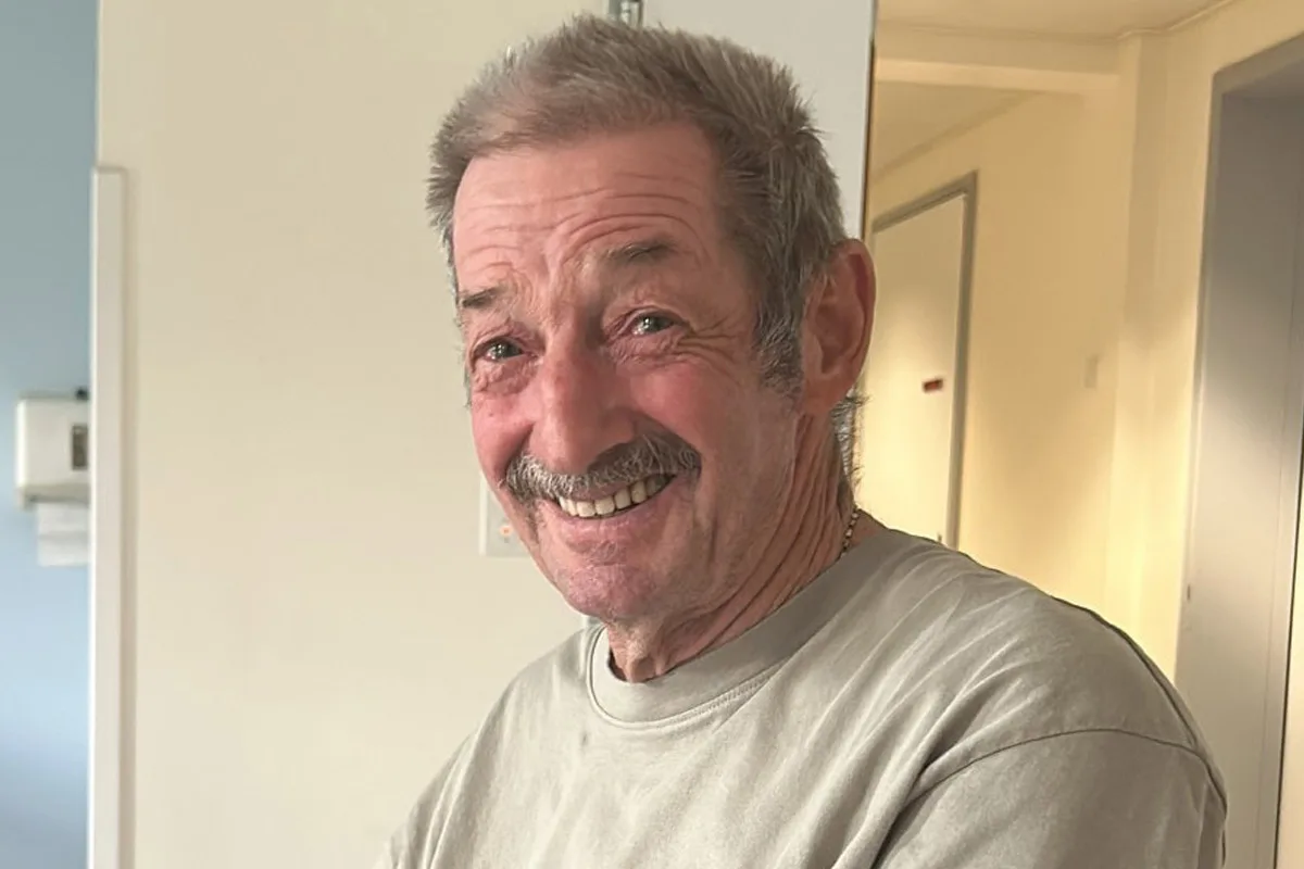 Ein älterer Mann mit einem Schnurrbart lächelt in die Kamera. Er befindet sich in einem hellen Raum mit weißen Wänden.