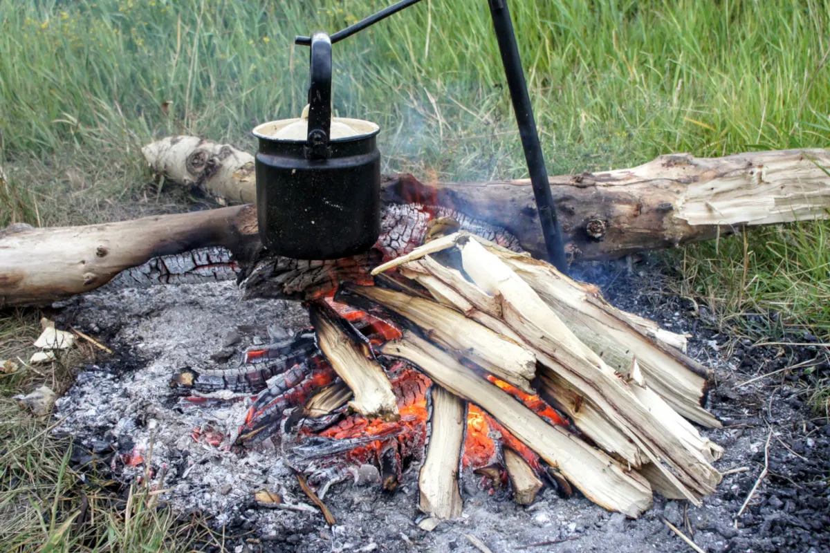Ein schwarzer Kessel hängt über einem offenen Lagerfeuer. Im Hintergrund ist Gras zu sehen.