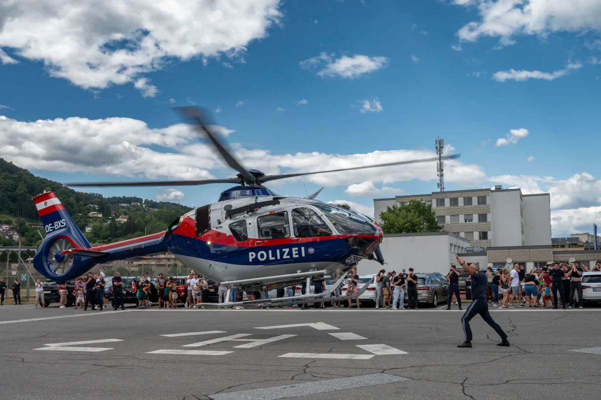 Ein Polizeihubschrauber schwebt über einem Parkplatz, während zahlreiche Menschen am Rand stehen und zuschauen. Im Hintergrund sind Gebäude und Landschaft zu sehen.