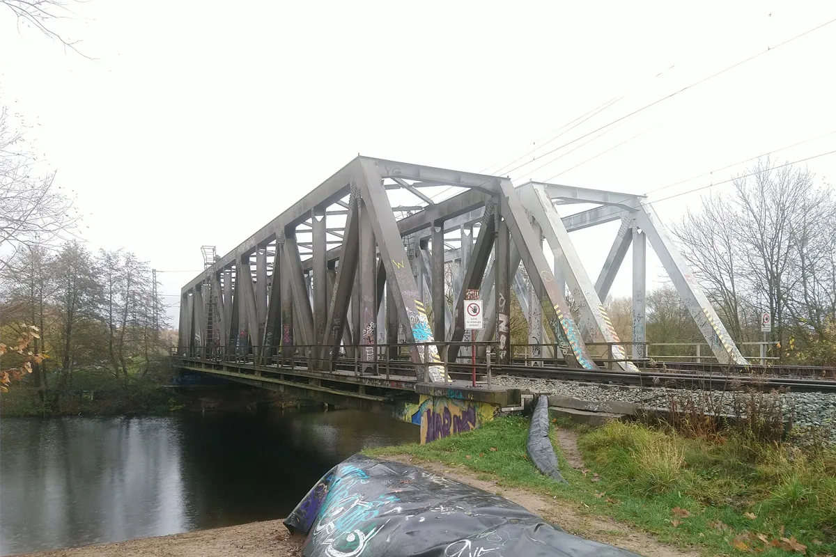 Eine Metallbrücke, die mit Graffiti bedeckt ist, erstreckt sich über einen ruhigen Fluss. Die Szene ist von herbstlicher Vegetation umgeben.