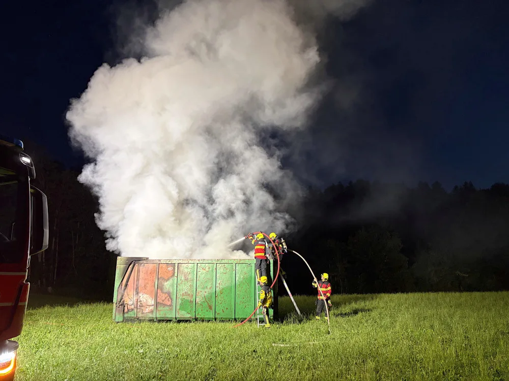 Nachts löschen Feuerwehrleute mit Schläuchen einen rauchenden Container auf einem Feld. Dicker Rauch steigt auf.