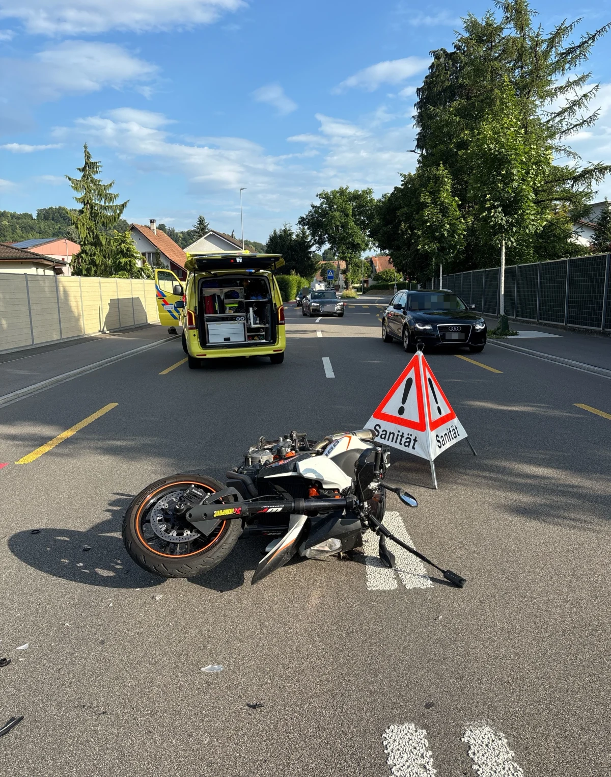 Ein Motorrad liegt umgestürzt auf einer Straße, während im Hintergrund ein Rettungswagen mit geöffneten Türen steht.