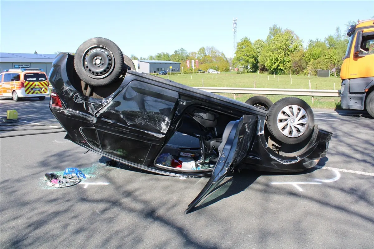 Ein schwarzes Auto ist auf dem Dach auf der Straße umgekippt. Daneben sind Trümmer und ein Lastwagen zu sehen. Es ist tagsüber, mit einem grünen Hintergrund aus Bäumen.