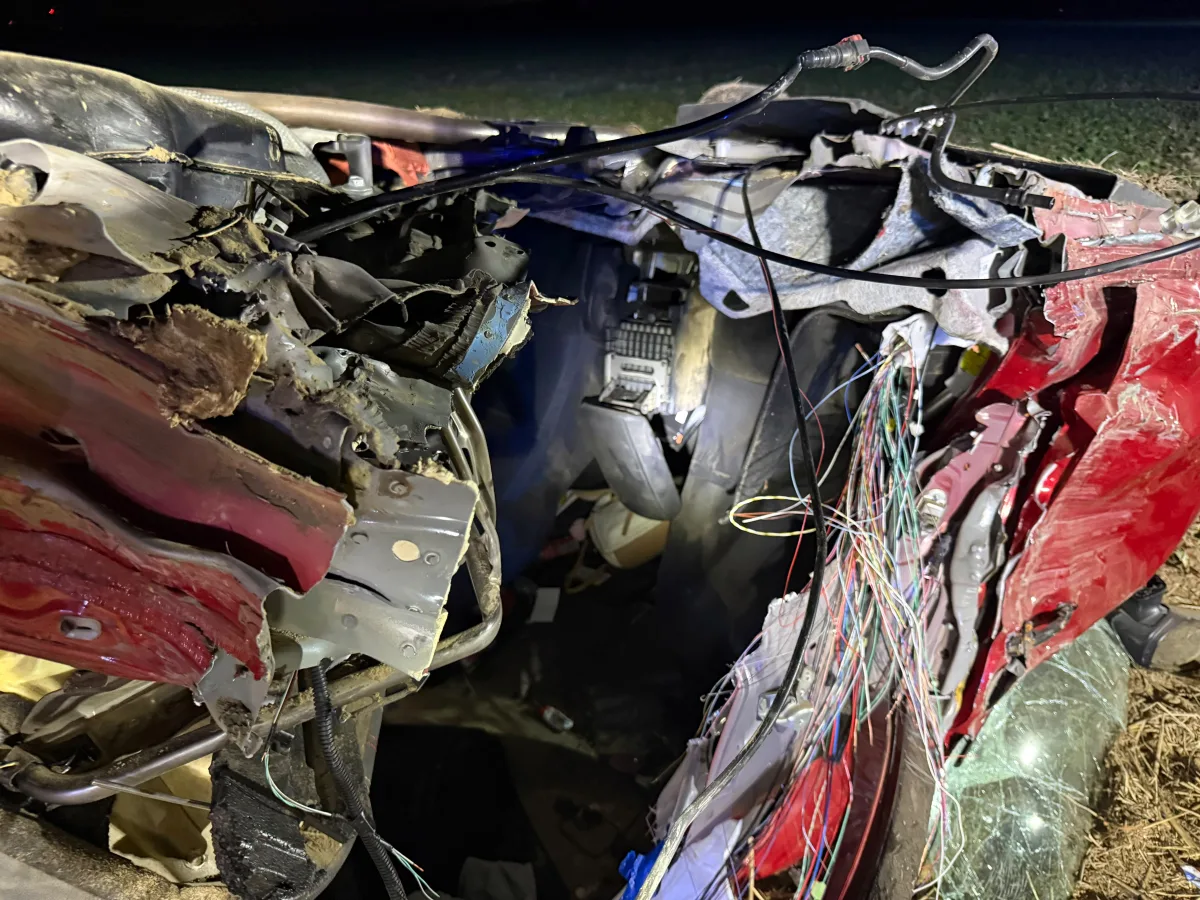 Nahaufnahme eines stark beschädigten roten Fahrzeugwracks bei Nacht. Sichtbare freiliegende Kabel, zerfetztes Metall und zerstörte Fahrzeugteile.