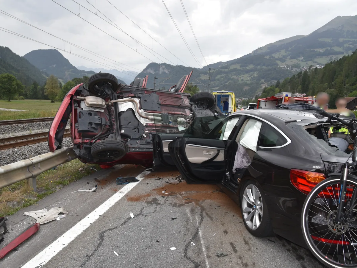 Ein roter Van ist umgekippt und liegt auf der Straße, während ein schwarzer BMW beschädigt daneben steht. Im Hintergrund sind Berge und Notfallfahrzeuge zu sehen.