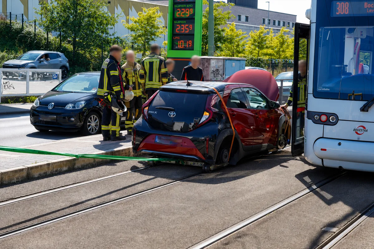 Ein roter Toyota Aygo steht beschädigt auf einer Straßenbahnspur. Feuerwehrleute sind vor Ort. Ein Bus ist daneben sichtbar.