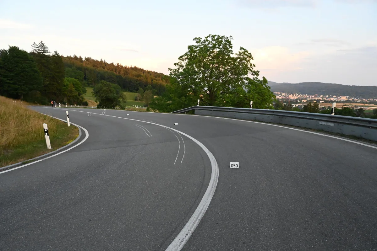 Eine Asphaltstraße mit Kurve, Fahrbahnmarkierungen und einer Leitplanke, umgeben von Bäumen.