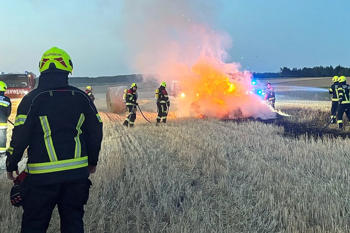 Mehrere Feuerwehrleute sind dabei, ein brennendes Fahrzeug auf einem Feld zu löschen. Dicker Rauch steigt auf und die Umgebung ist abgeerntet.