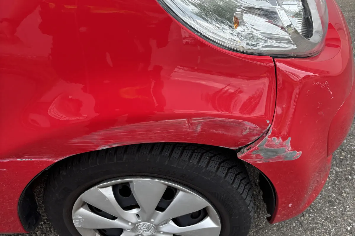 Ein rotes Auto zeigt deutliche Kratzer und Beulen am vorderen rechten Kotflügel. Der Reifen ist ebenfalls sichtbar.