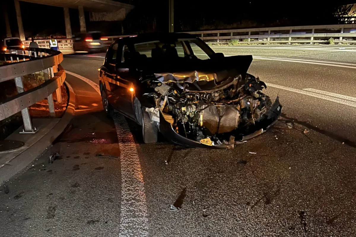 Das Bild zeigt ein Auto mit schwer beschädigter Front auf einer Straße bei Nacht. Im Hintergrund sind weitere Fahrzeuge und eine Leitplanke zu sehen.