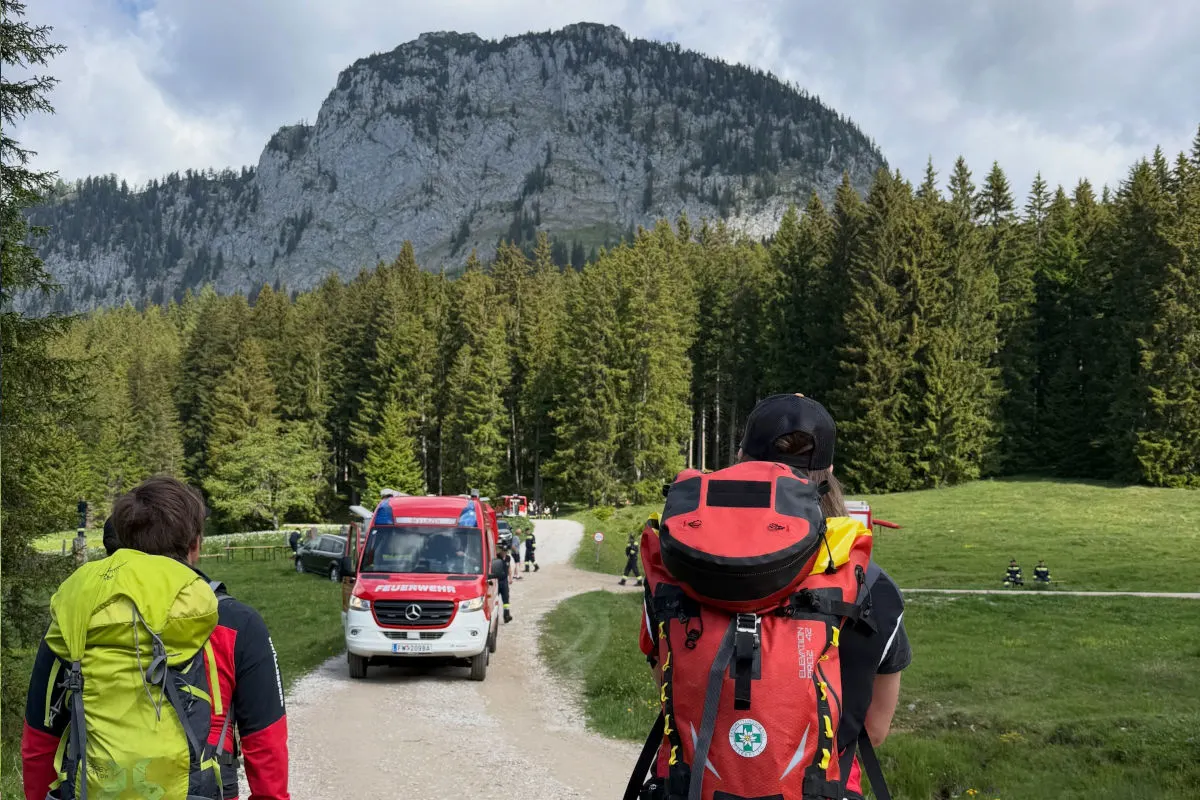 Auf einem Waldweg steht ein Feuerwehrfahrzeug. Zwei Personen mit Wanderrucksäcken blicken in Richtung des Fahrzeugs, umgeben von Bäumen und einem felsigen Berg im Hintergrund.