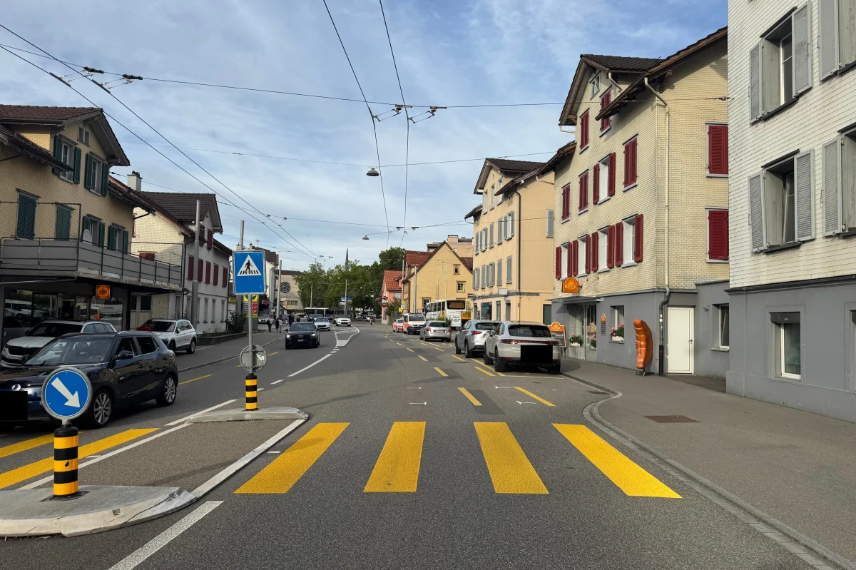 Eine Straße in der Stadt mit gelben Zebrastreifen, parkenden Autos und einigen Geschäften im Hintergrund. Fußgängerüberweg und Straßenschilder sind sichtbar.