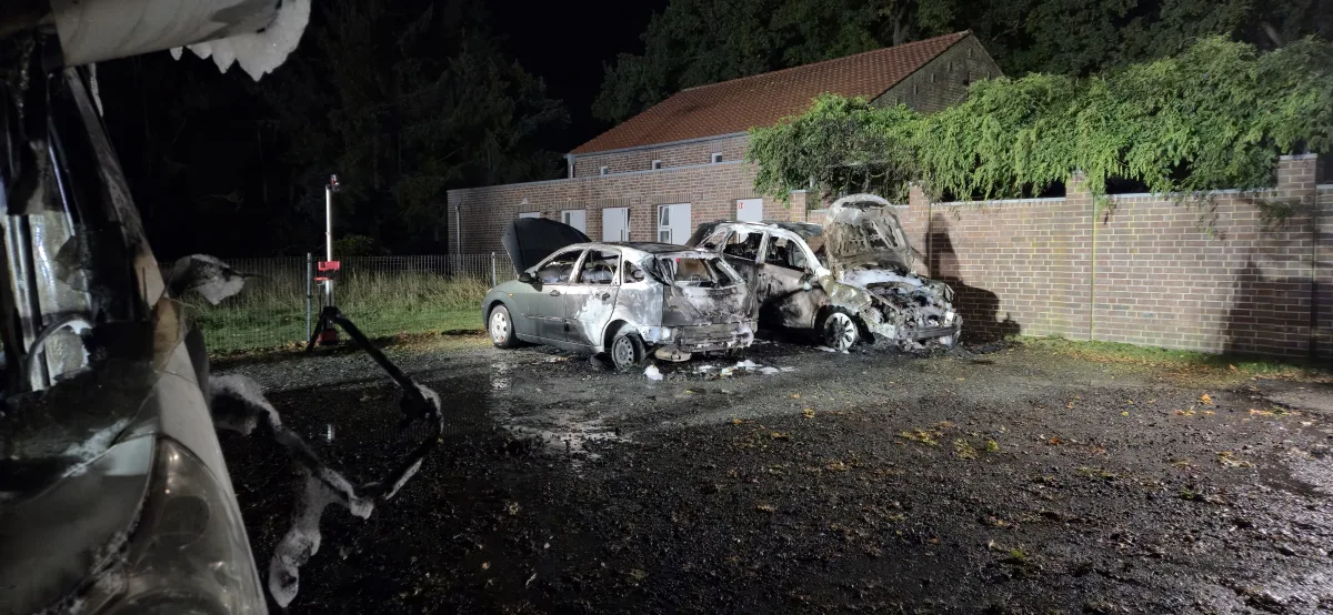 Zwei ausgebrannte Autos stehen nebeneinander auf einem nächtlichen Parkplatz; verkohlte Überreste sind sichtbar.