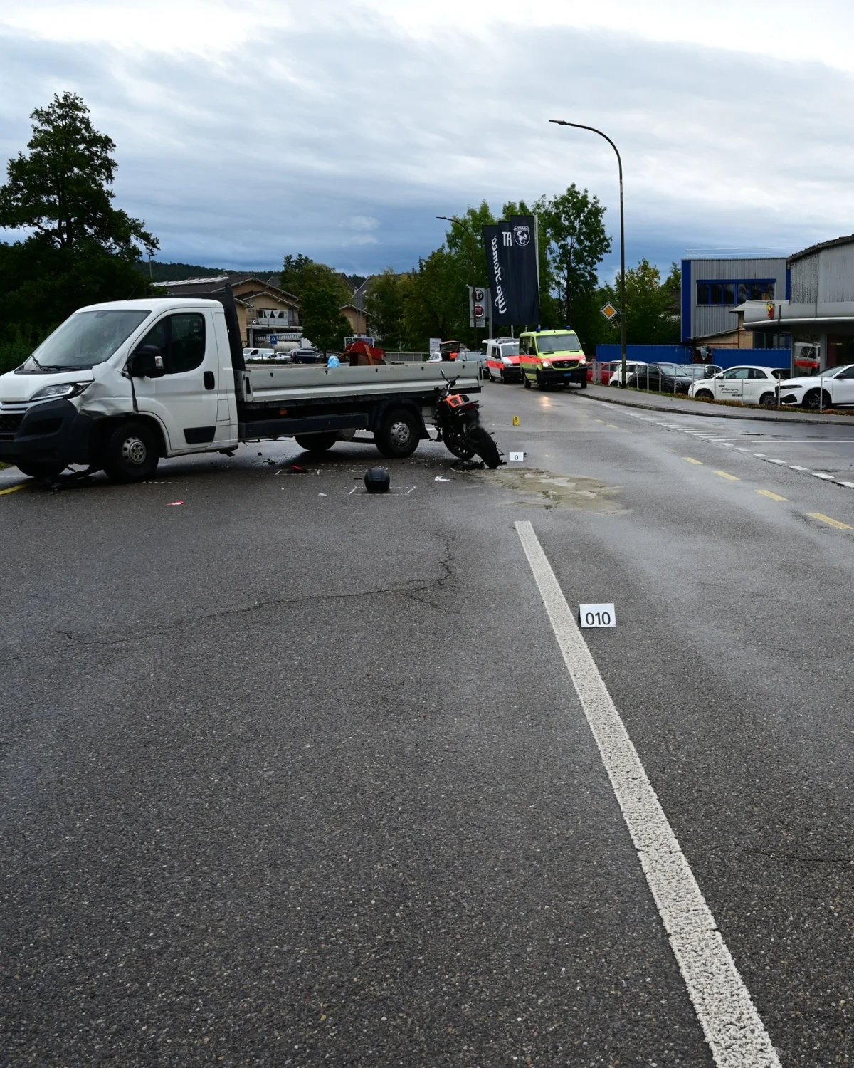 Ein beschädigtes Motorrad liegt neben einem weißen Lieferwagen auf der nassen Straße. Im Hintergrund steht ein Rettungswagen.
