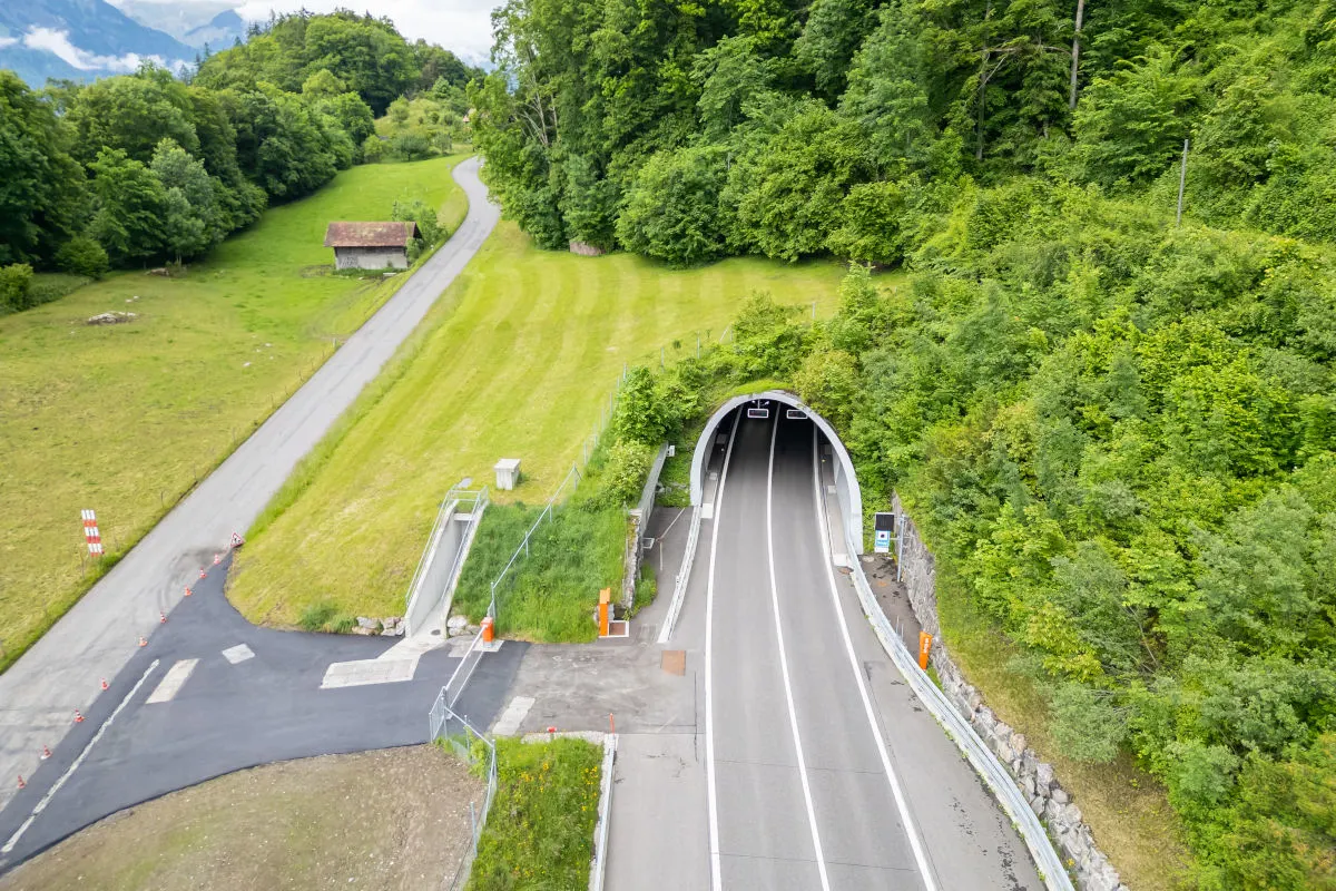 Ein Straßentunnel führt durch eine grüne, bewaldete Landschaft in bergigem Gebiet. Ein kleiner Weg verläuft neben einer Wiese.