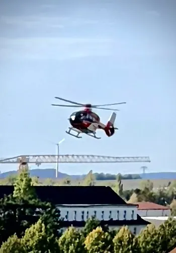 Ein roter Rettungshubschrauber nähert sich einem Stadtgebiet. Im Hintergrund sind Gebäude und eine Windkraftanlage sichtbar.