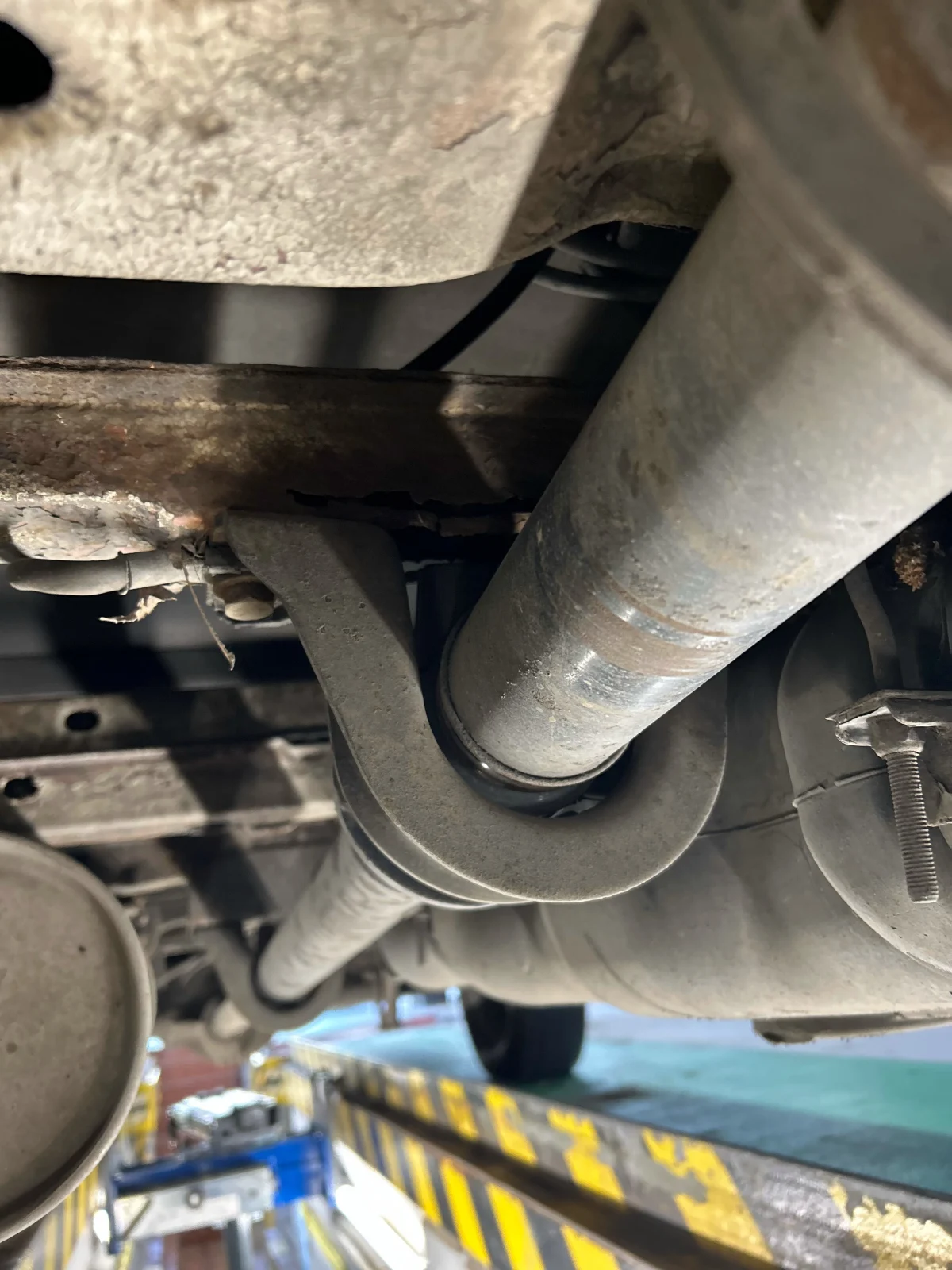 Ein Bild der Unterseite eines Fahrzeugs zeigt ein Auspuffsystem, das über einer Werkstattgrube verläuft. Verschiedene Fahrzeugteile sind sichtbar, darunter Rohre und Halterungen.