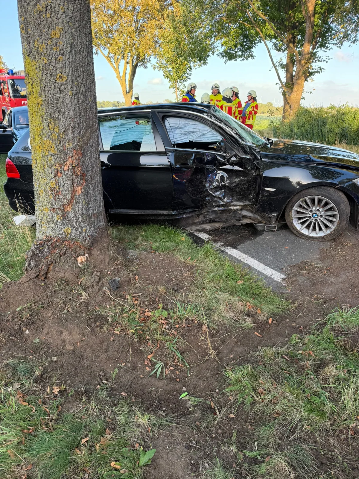 Ein schwarzes Auto mit Schäden an der linken Seite steht nahe einem Baum. Feuerwehrleute in gelben Schutzanzügen stehen im Hintergrund auf einer Straße.