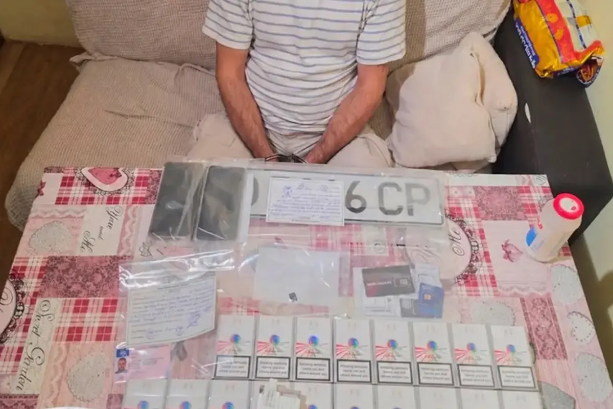 Eine Person in gestreiftem T-Shirt und Handschellen sitzt hinter einem Tisch, der mit Zigarettenpackungen, Dokumenten und Kennzeichen bedeckt ist.