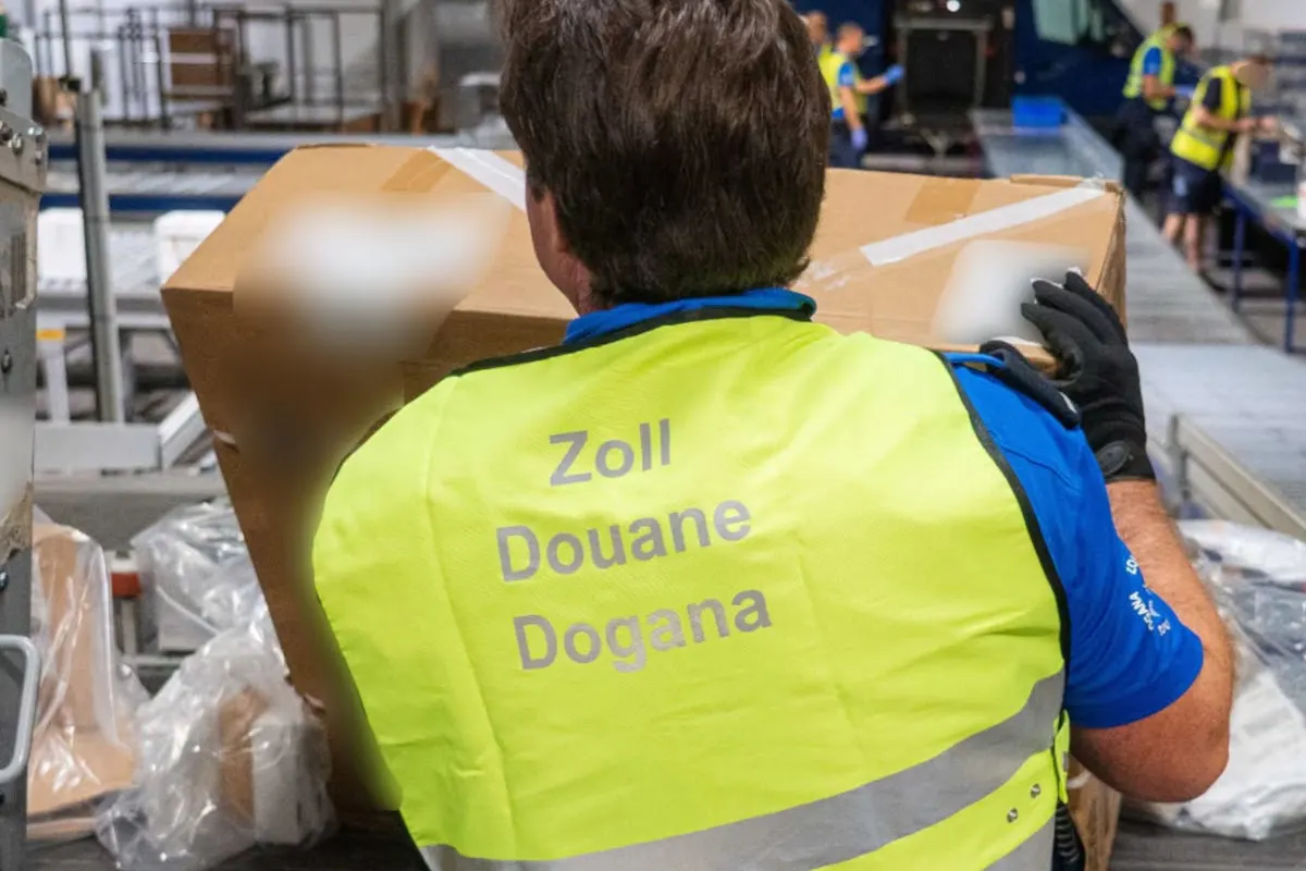 Ein Zollbeamter in gelber Weste hebt ein Paket in einer Lager- oder Verteilhalle an. Im Hintergrund sind weitere Beamte bei der Arbeit.