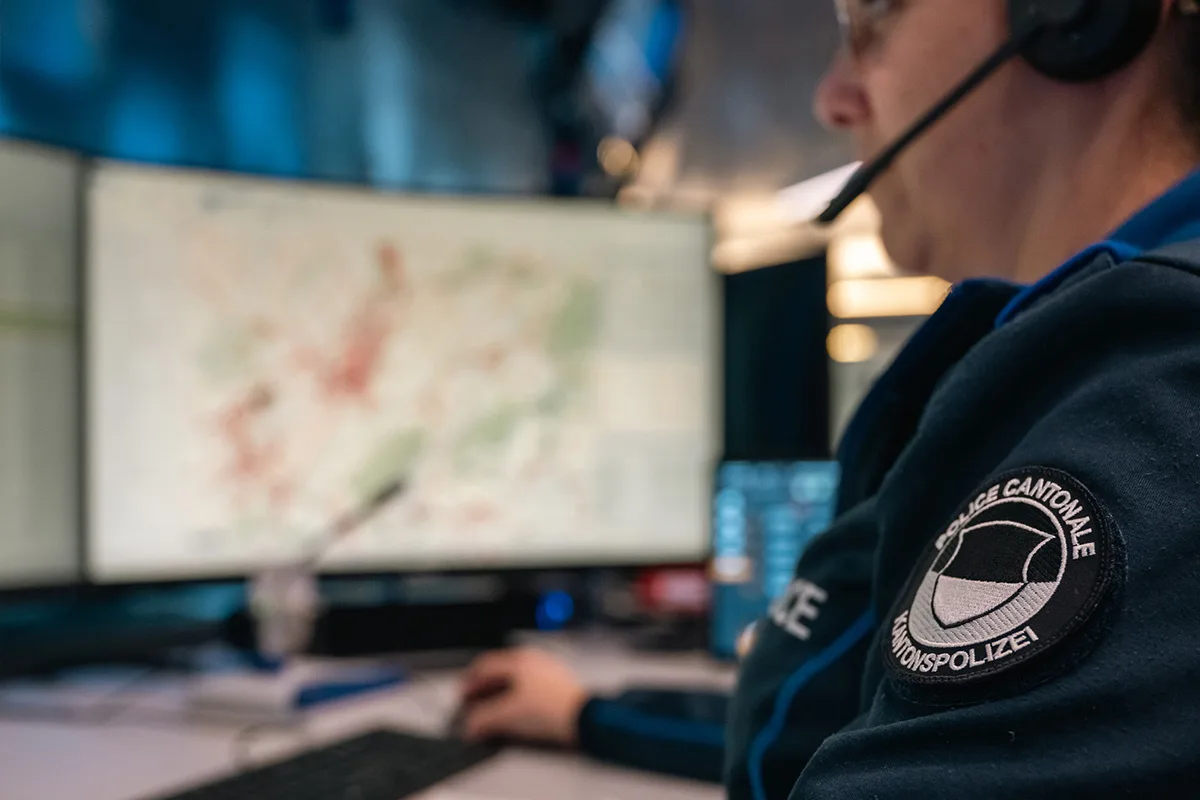 Eine Polizistin in einer Einsatzzentrale arbeitet konzentriert vor einem großen Bildschirm. Auf ihrer Uniform ist das Wappen der Kantonspolizei sichtbar.