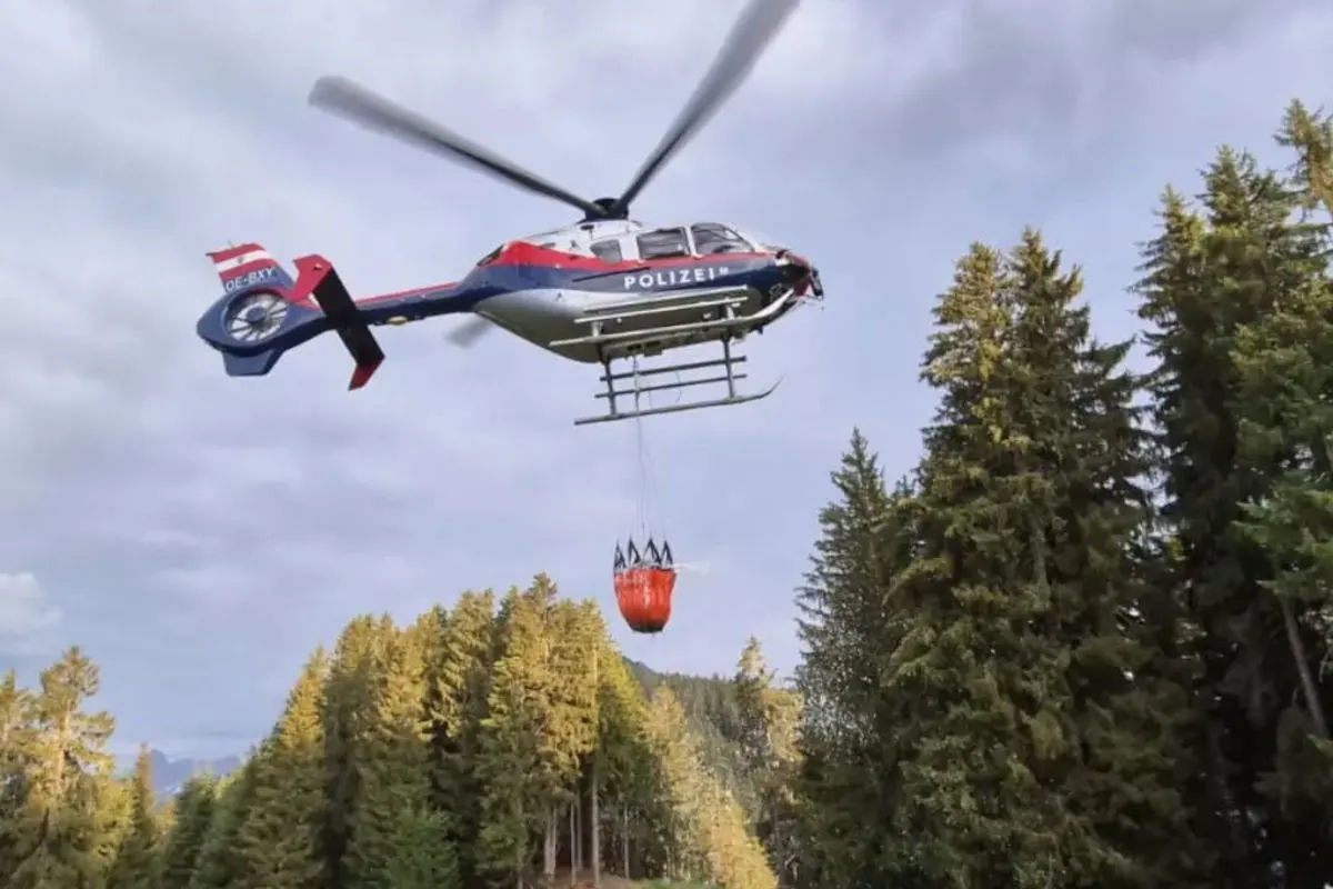 Ein Polizeihelikopter transportiert einen roten Löschbehälter und fliegt über einen dichten Nadelwald. Der Himmel ist bewölkt.