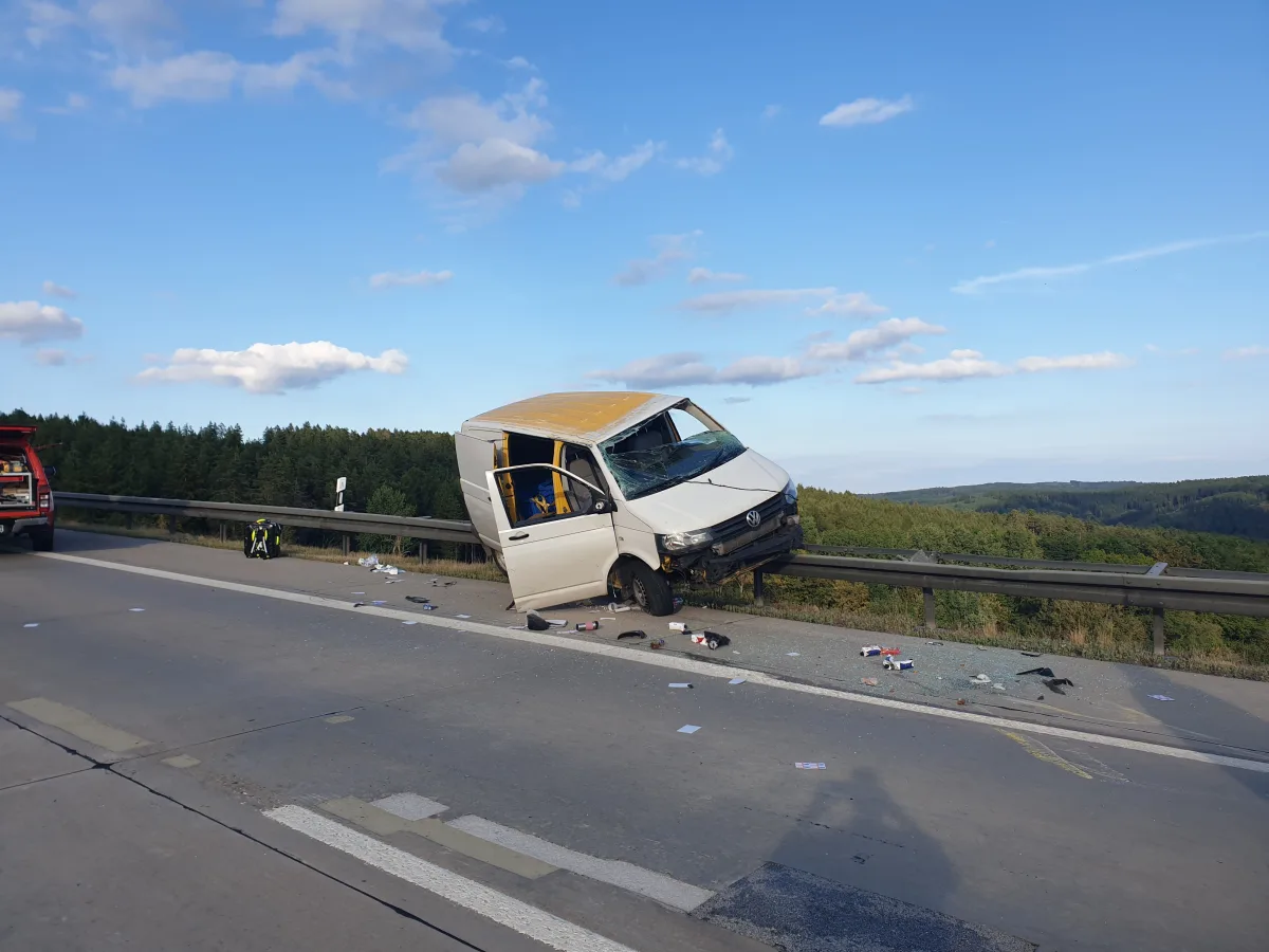 Ein weißer VW-Transporter mit gelbem Dach lehnt beschädigt gegen eine Leitplanke auf einer Autobahn. Auf der Fahrbahn liegen Trümmer und Glassplitter.