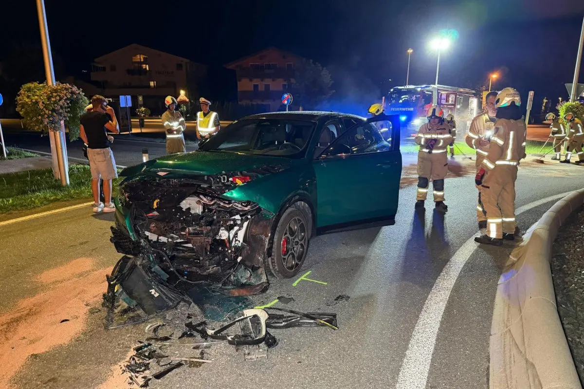 Ein grünes Auto mit stark beschädigter Front steht auf der Straße, umgeben von Feuerwehrleuten in Uniformen bei Nacht.