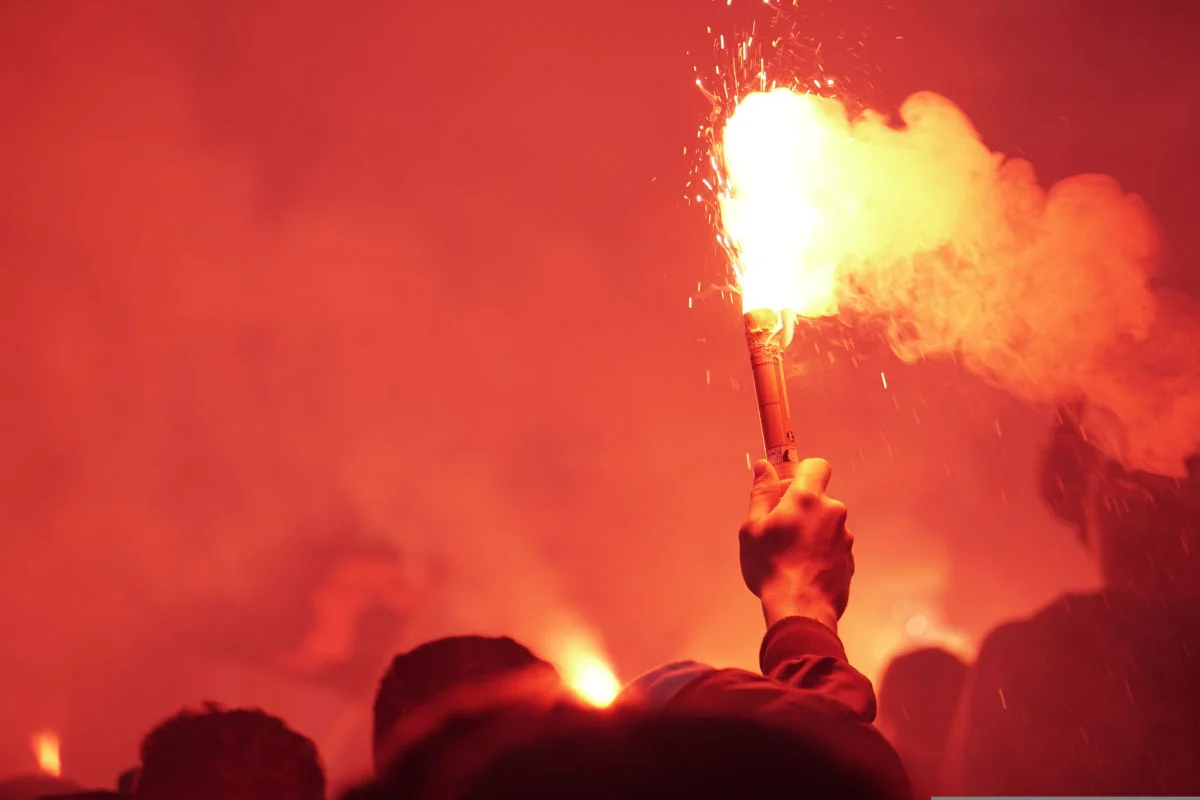 In einer dicht gedrängten Menschenmenge hält jemand eine helle, leuchtende rote Fackel nach oben. Dampf oder Rauch umgibt die Flamme.