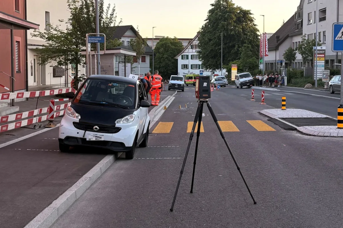 Auf dem Bild steht ein weiß-schwarzer Smart quer auf einem Gehweg in einer Wohnstraße. Im Hintergrund sind Personen in Sicherheitskleidung und ein Vermessungsgerät zu sehen.