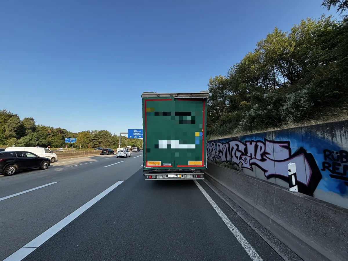Ein Lastwagen fährt auf der rechten Spur einer Autobahn, umgeben von weiteren Fahrzeugen. Am Straßenrand ist Graffiti zu sehen.
