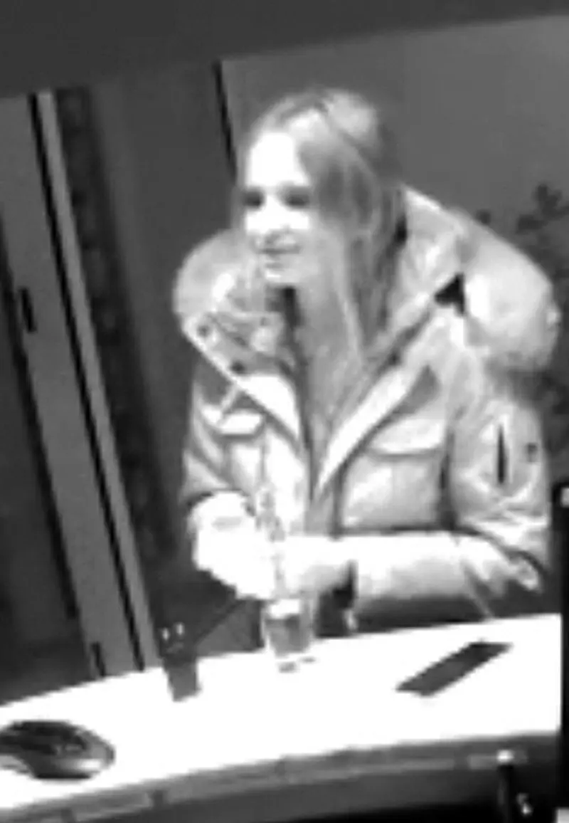 Eine Frau in einer Winterjacke steht mit einem Getränk in der Hand an einem Tresen in einer Bar. Das Bild ist schwarz-weiß.