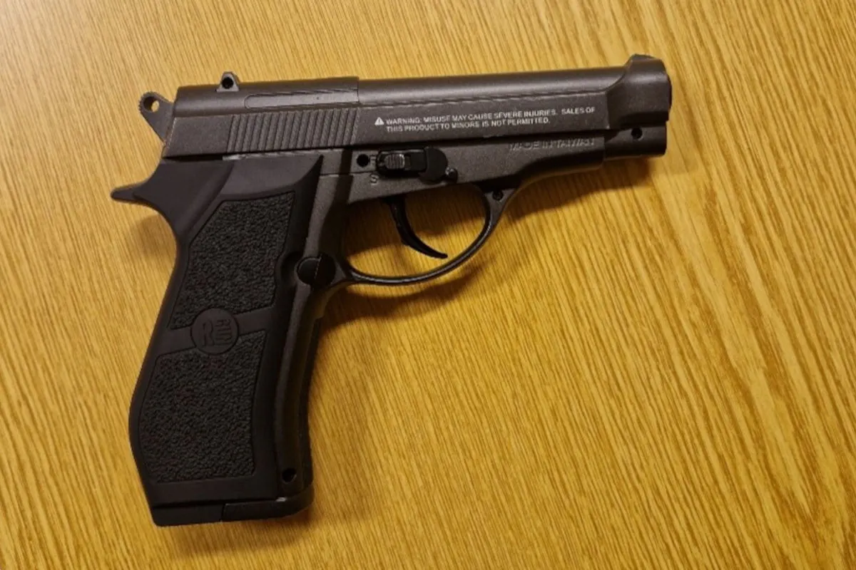 Auf dem Bild ist eine schwarze Pistole zu sehen, die auf einem Holztisch liegt. Die Waffe ist detailliert abgebildet.