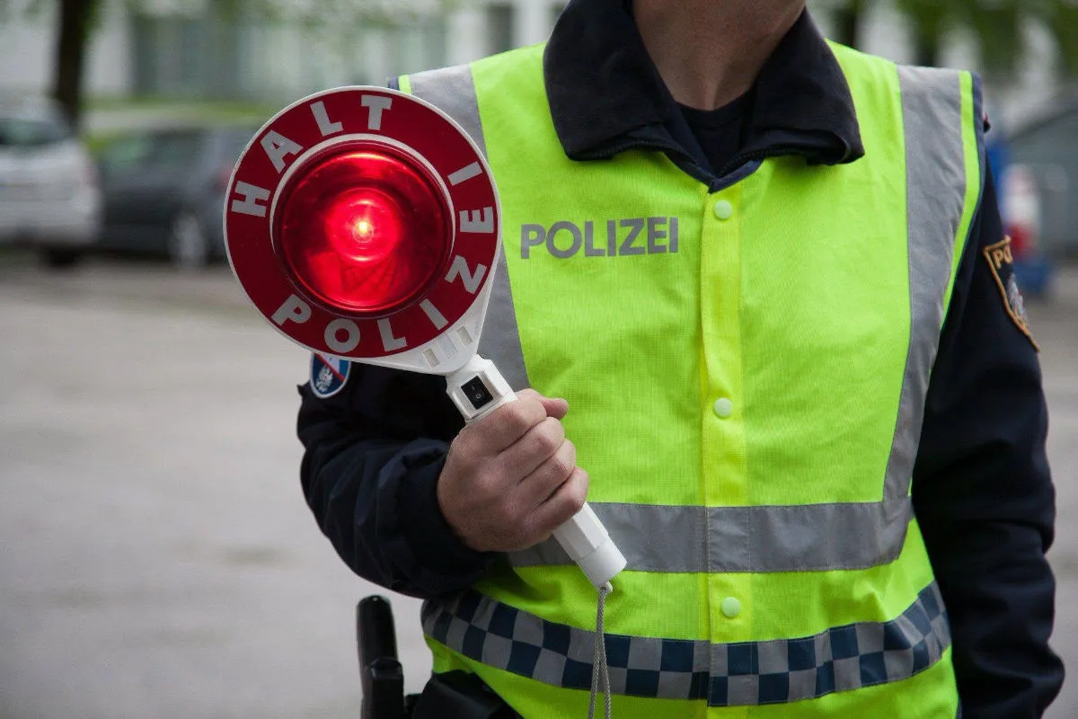 Ein Polizist in einer gelben Warnweste hält eine beleuchtete Polizeikelle mit der Aufschrift 'Halt Polizei', umgeben von Fahrzeugen auf einer Straße.