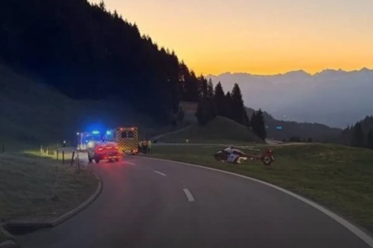 Ein Rettungshubschrauber steht auf einem Feld, daneben sind Einsatzfahrzeuge mit eingeschaltetem Blaulicht auf einer kurvigen Bergstraße sichtbar. Der Himmel zeigt einen Sonnenaufgang über den Bergen.