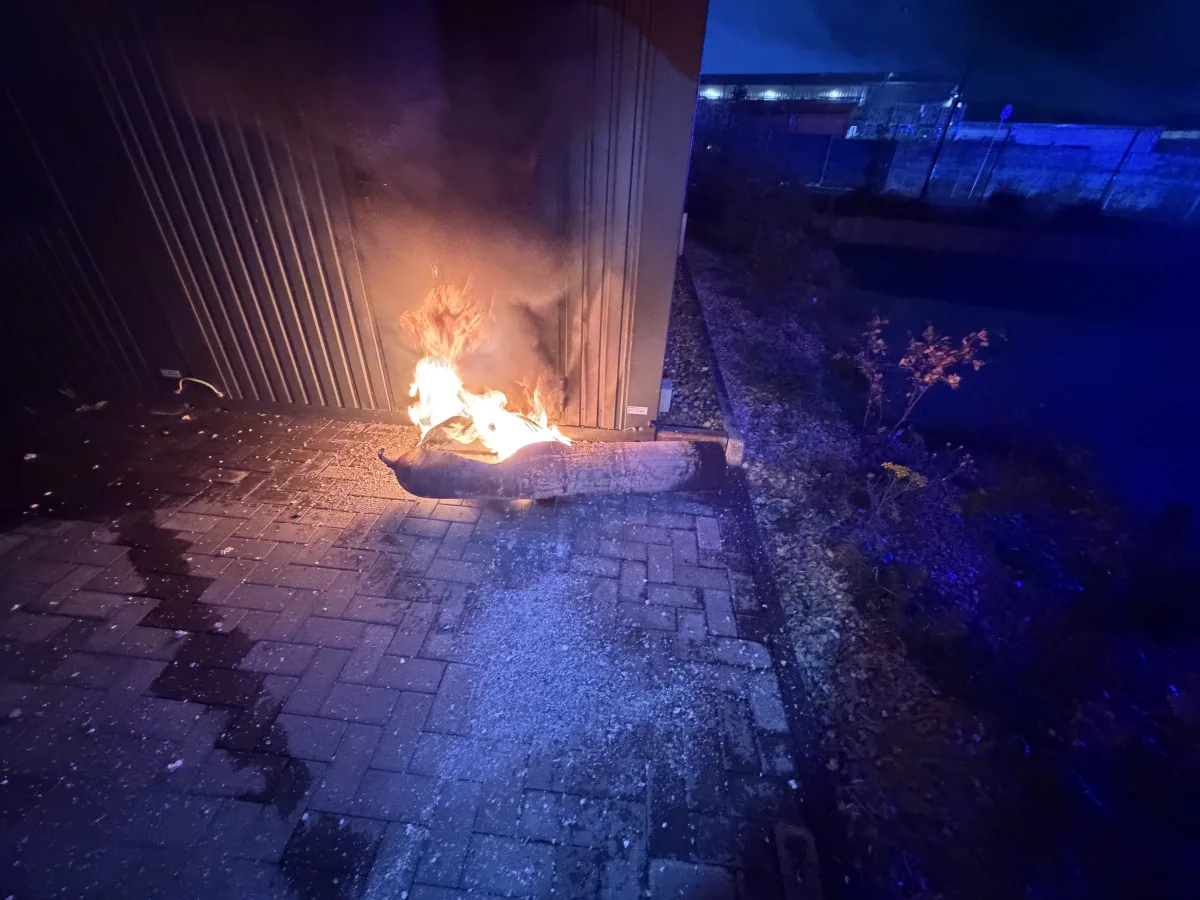 Ein längliches Objekt brennt vor einer metallenen Gebäudewand auf einem gepflasterten Weg bei Nacht.